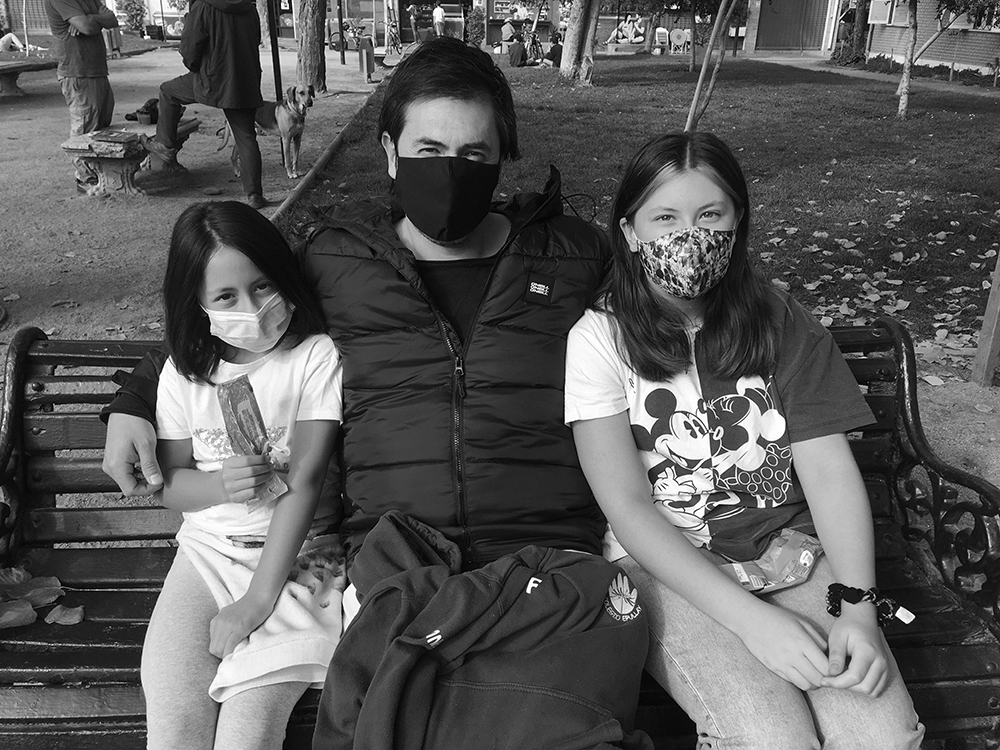 Un hombre y dos niñas con mascarilla, sentados en un escaño de plaza durante la pandemia
